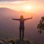 Une jeune femme, les bras tendus, se tient sur le flanc d'une colline et regarde le soleil couchant.
